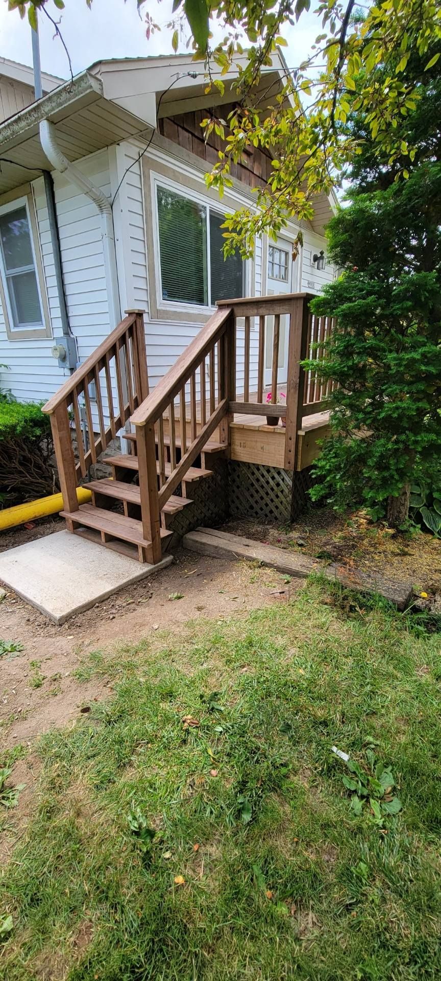 A white house with wood stairs leading up to a small wooden deck, positioned next to a green tree and a lawn.