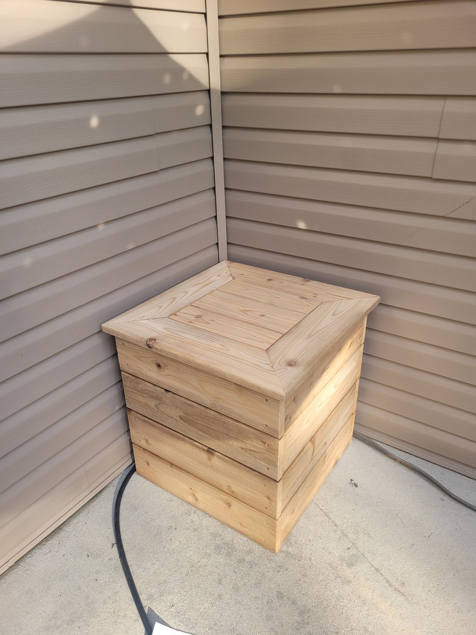 A square wooden box with a lid sits in the corner of a patio against brown siding walls.