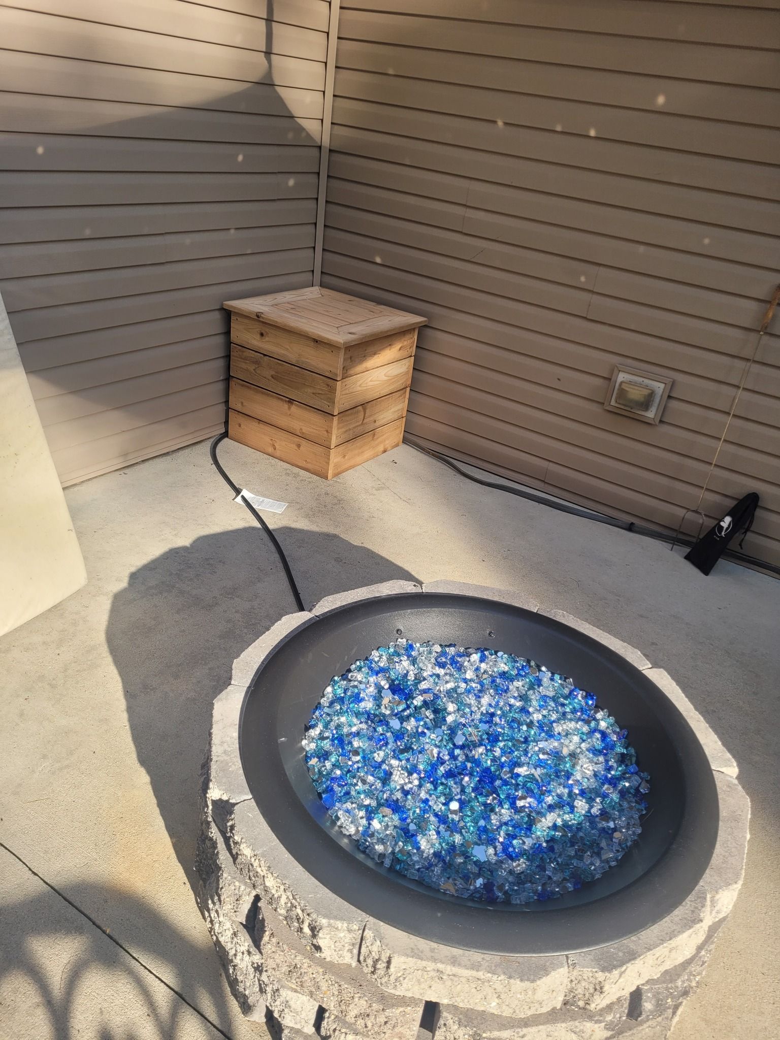 A circular concrete fire pit filled with blue and clear glass rocks, with a gas line connecting to a small wooden enclosure.