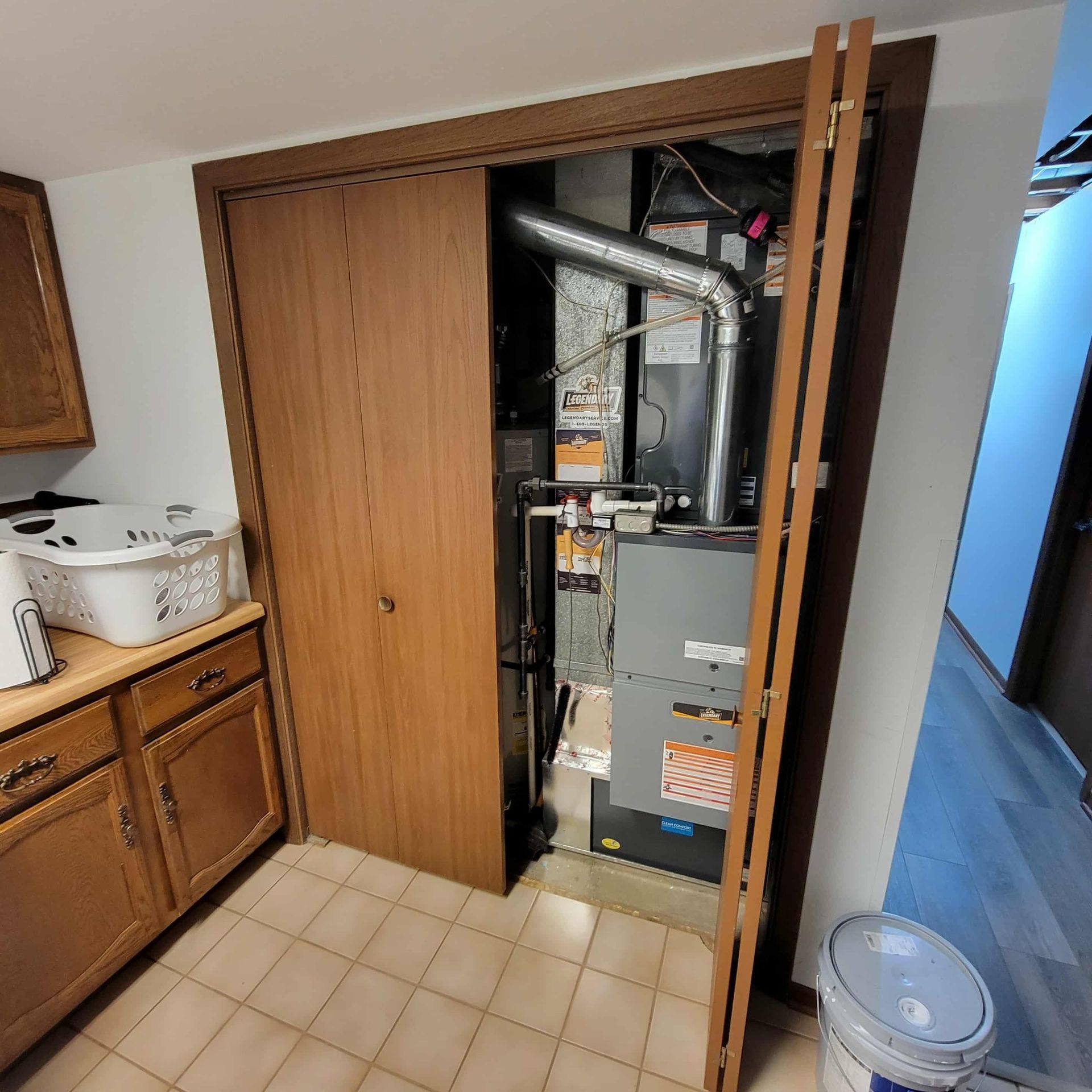 A closet containing a furnace and HVAC equipment sits next to wooden kitchen cabinets and a tiled floor.