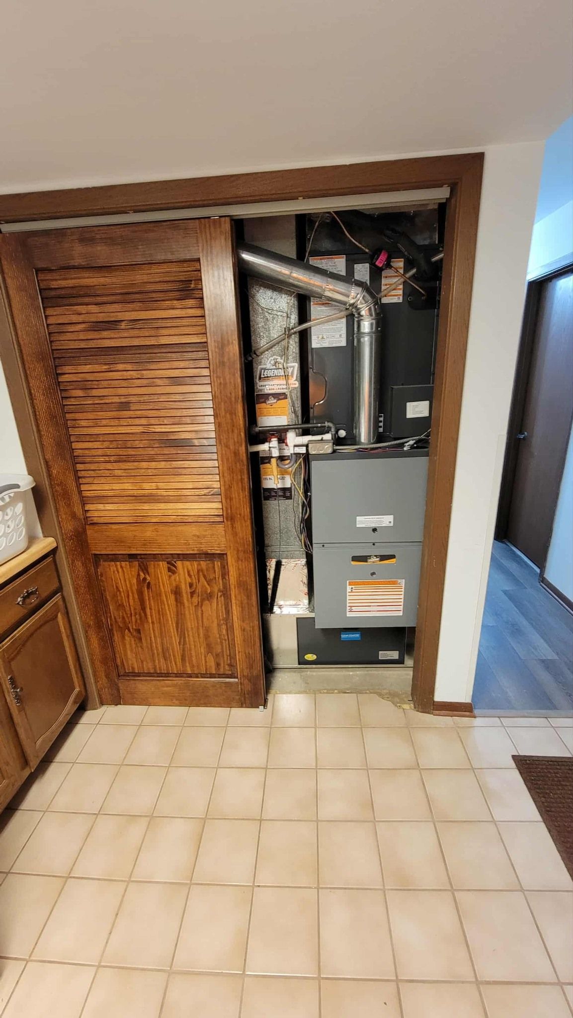 A gray furnace unit with exposed metal ductwork located inside an open closet with a wooden louvered door in a tiled room.