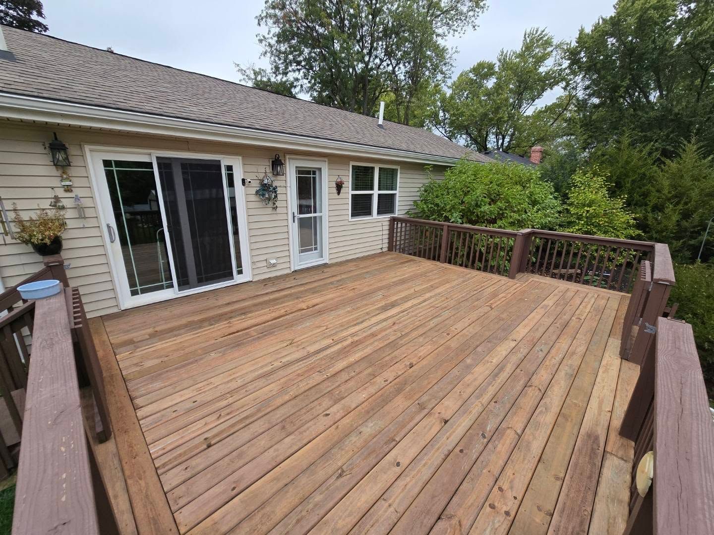 A wide, natural-toned wooden deck attached to a light-sided house with a sliding glass door and windows.