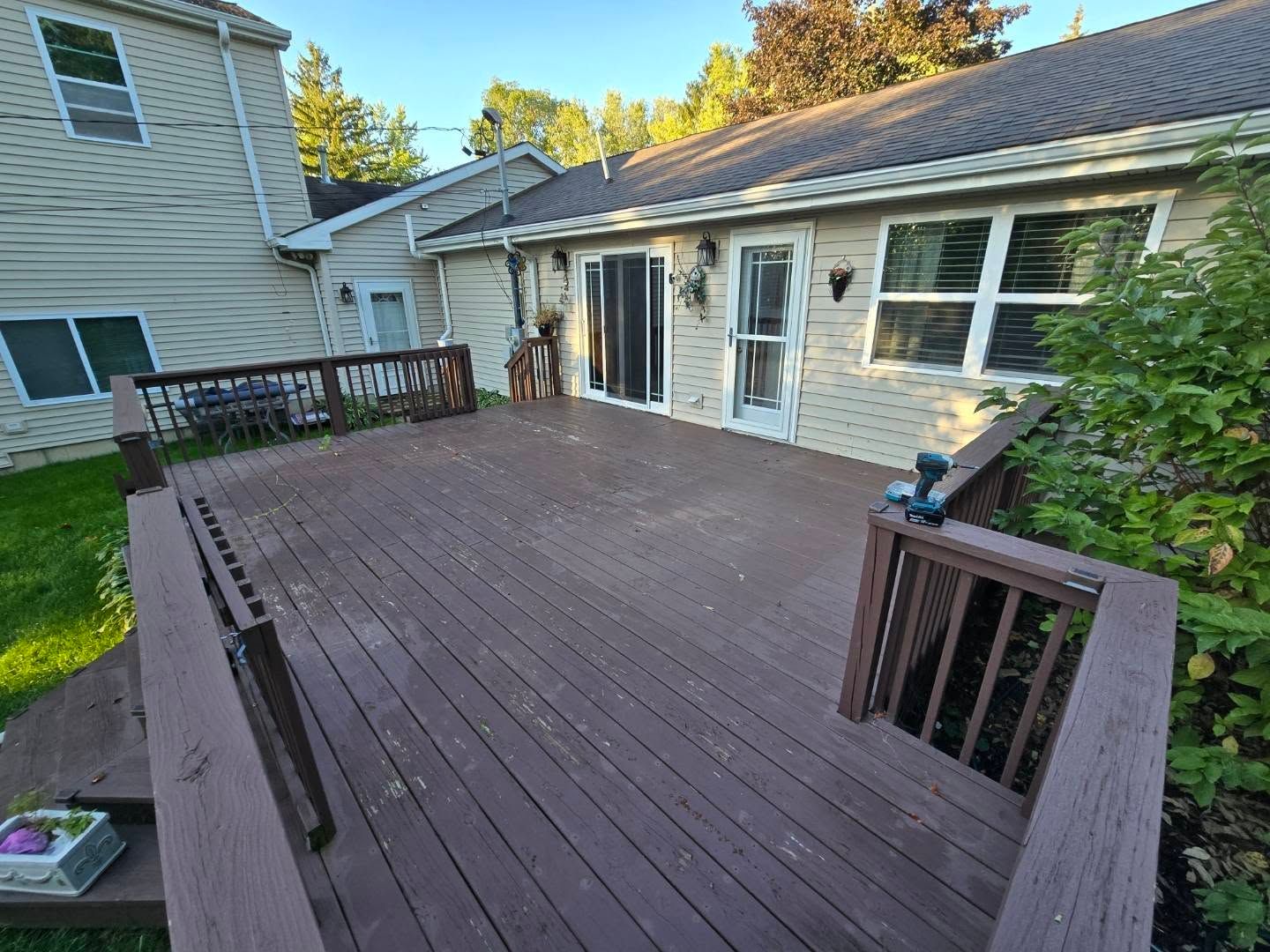A large, brown, elevated wooden deck attached to a light-colored house with green trees in the background.