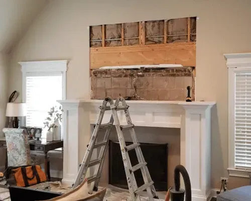 A silver ladder stands before a fireplace with an open wall cavity above the mantle, revealing wood studs and cinder blocks.