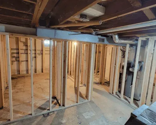Unfinished basement space showing new wooden wall framing, exposed ceiling joists, and installed HVAC ductwork.