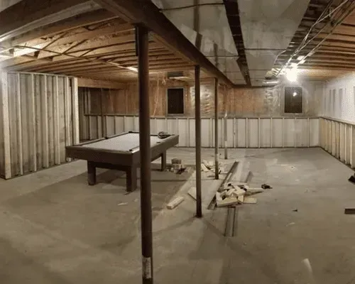 Unfinished basement with exposed ceiling joists, wood-framed walls, a pool table in the center, and concrete flooring.