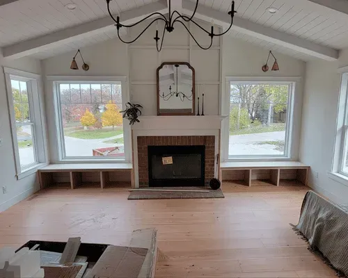 A bright living room with a brick fireplace, built-in window benches, hardwood floors, and a vaulted ceiling.