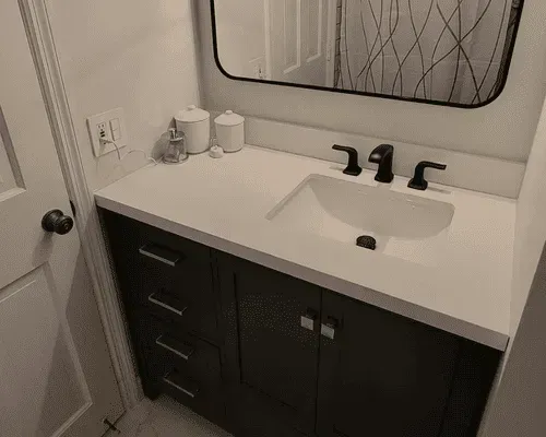 A modern bathroom vanity with a white countertop, dark cabinets, a black faucet, and a rectangular mirror.