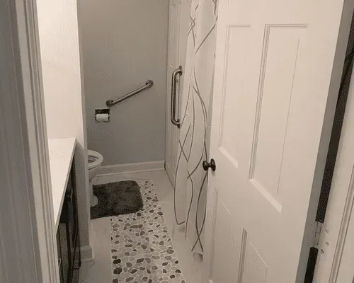 A bathroom view showing a toilet with a grab bar, a patterned rug on a white floor, and a white shower curtain to the right.