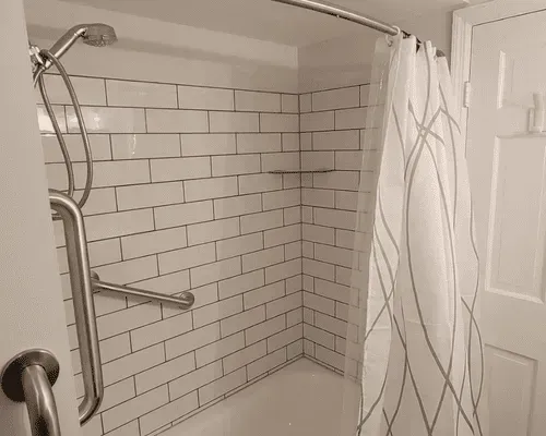 White subway tile shower with a handheld showerhead, grab bars, and a white patterned curtain.