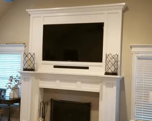 A white fireplace mantel featuring a built-in television, a soundbar, and two decorative metal lanterns on the ledge.