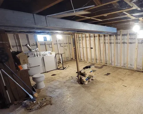 An unfinished basement with exposed wooden wall studs, a concrete floor, a central support post, and laundry appliances.