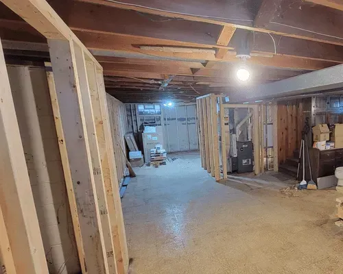 Unfinished basement with exposed ceiling beams, wood stud walls framing a future room, and a concrete floor.