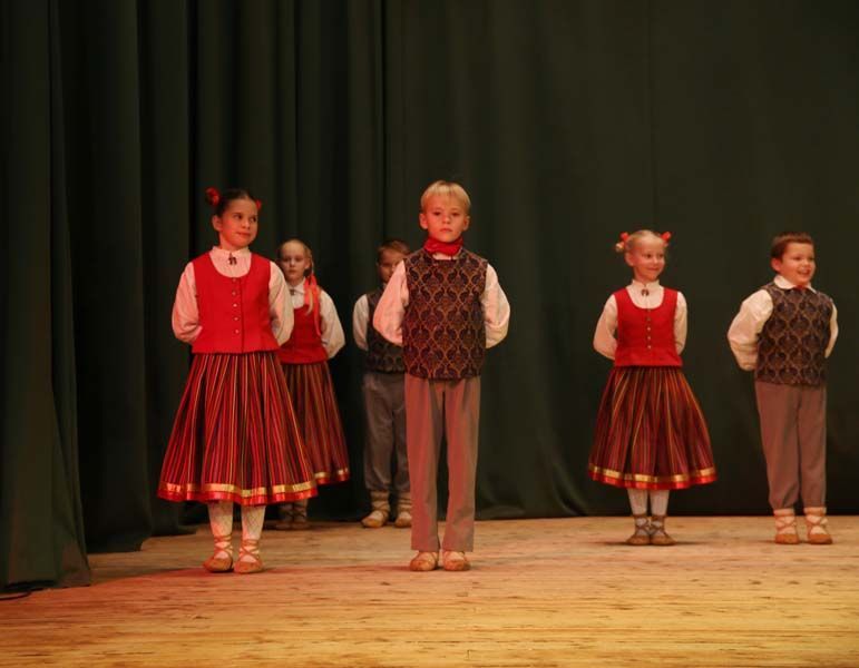 Bērnu grupa stāv uz skatuves sarkanās kleitās