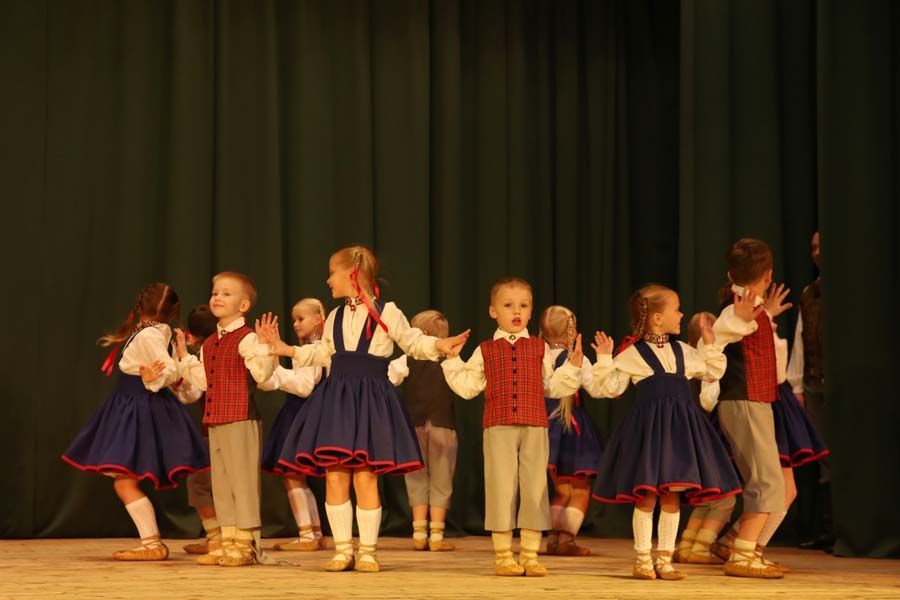 Bērnu grupa dejo uz skatuves zaļa aizkara priekšā.
