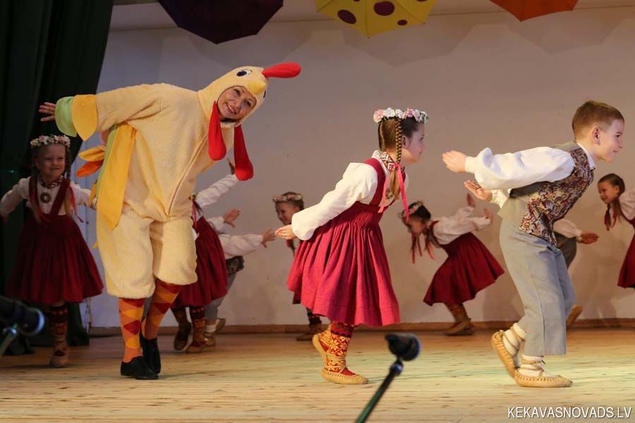 Bērnu grupa dejo uz skatuves kopā ar vīrieti vistas kostīmā.