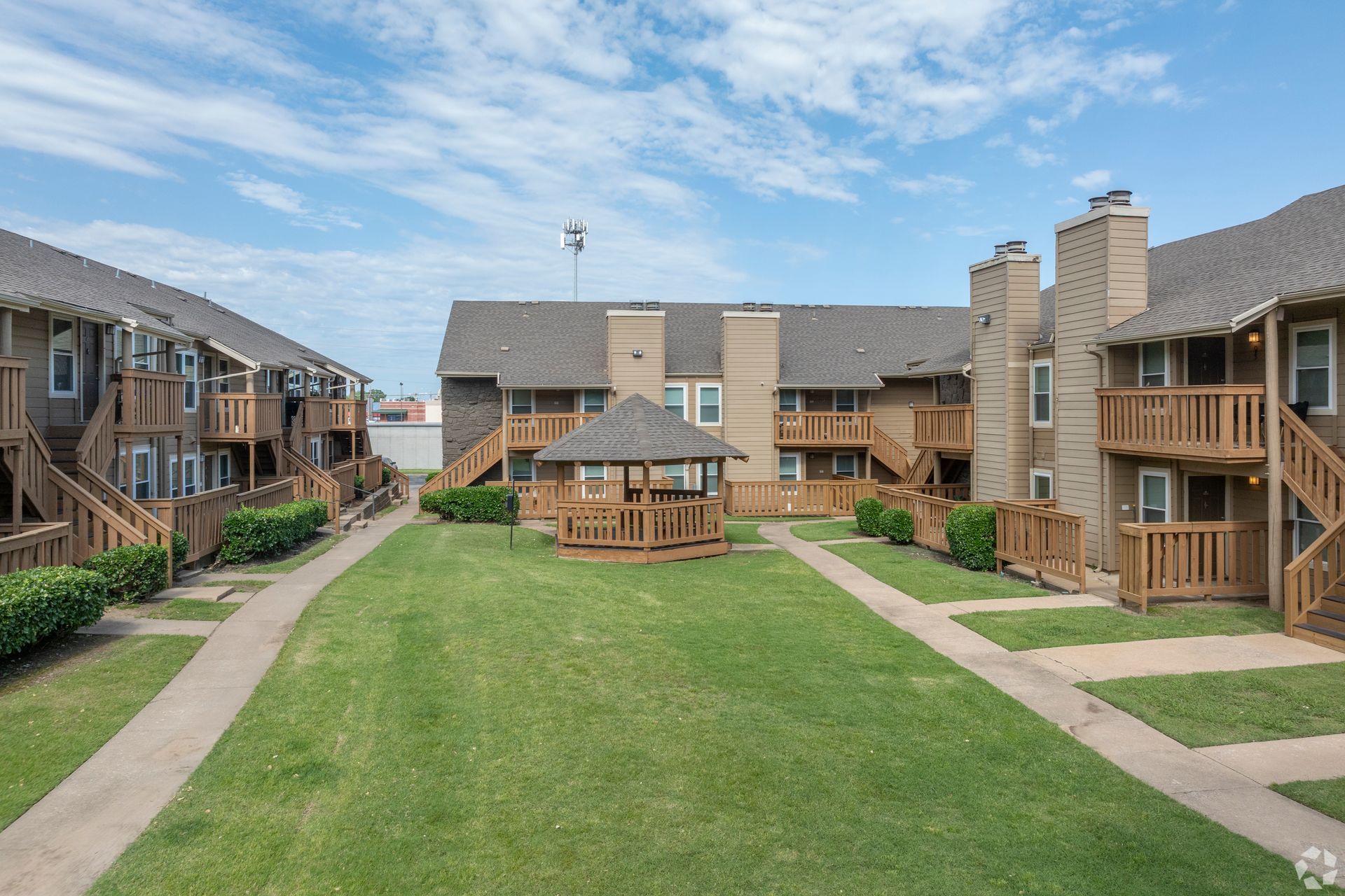 Apartment courtyard with green lawn, gazebo, and wooden balconies under a blue sky
