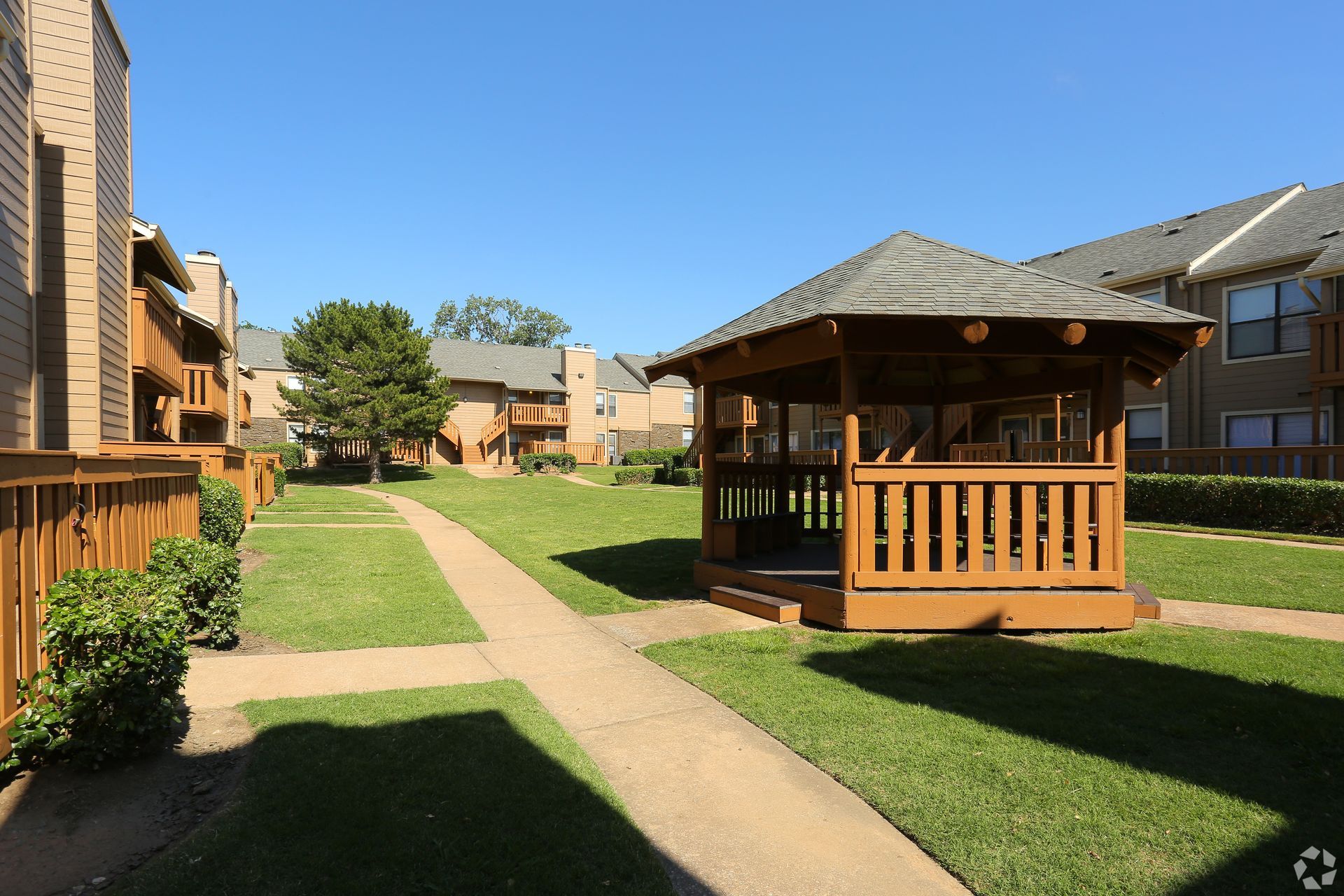 Apartment complex courtyard with a wooden gazebo, grassy lawn, and concrete walkway under a clear blue sky