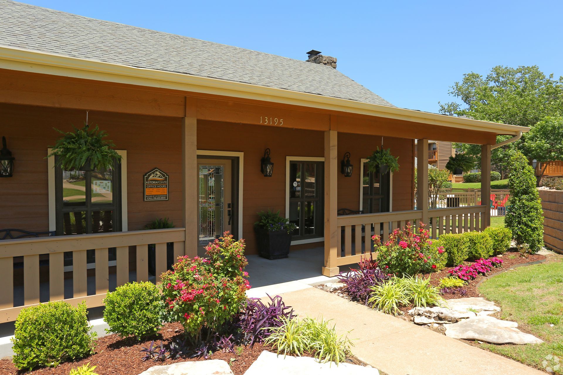Front porch of a tan house with potted plants, pink flowers, and a curved walkway in bright sunlight