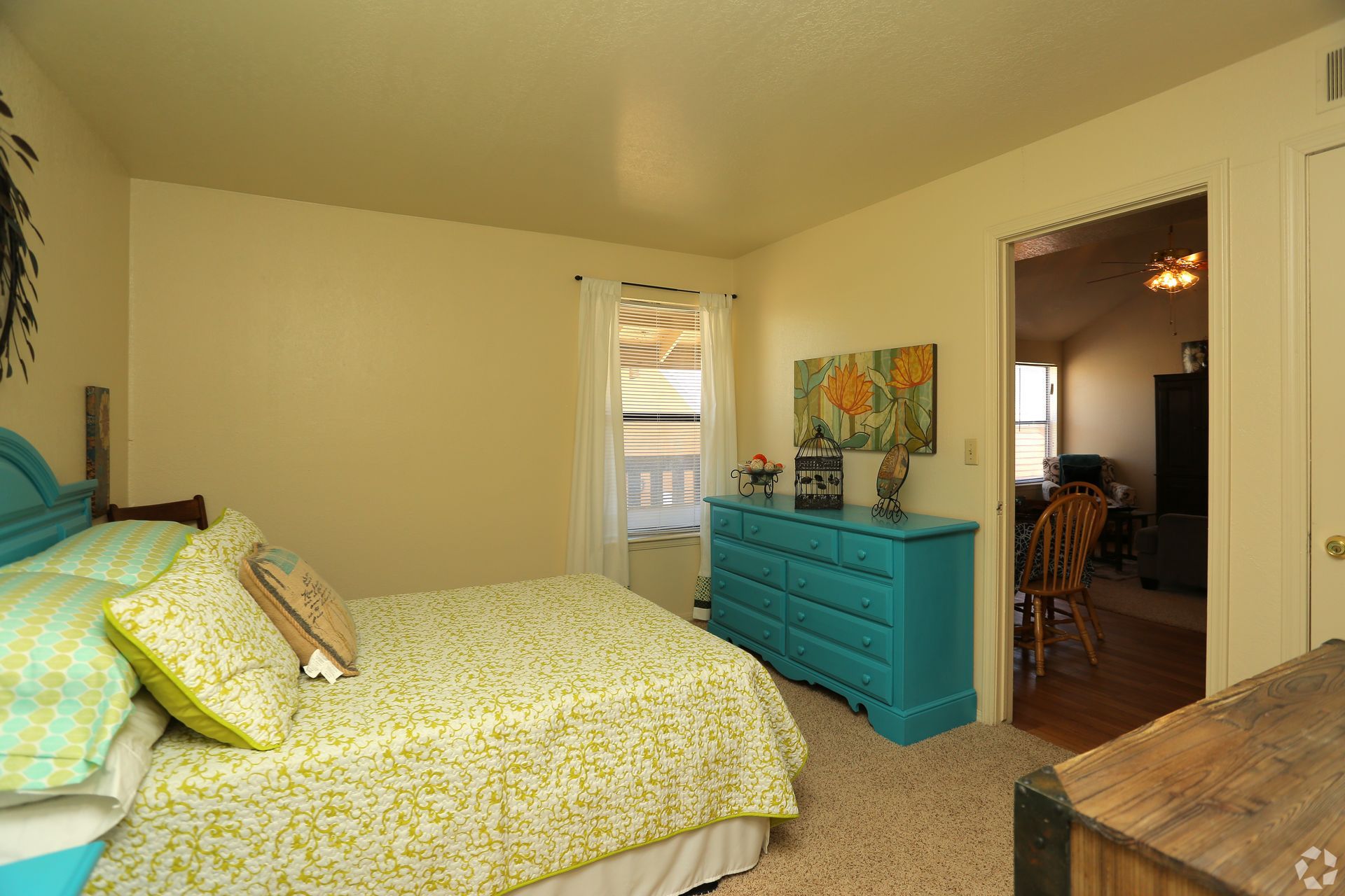 Bedroom with bed, turquoise dresser, and doorway to a dining area, in warm beige tones.