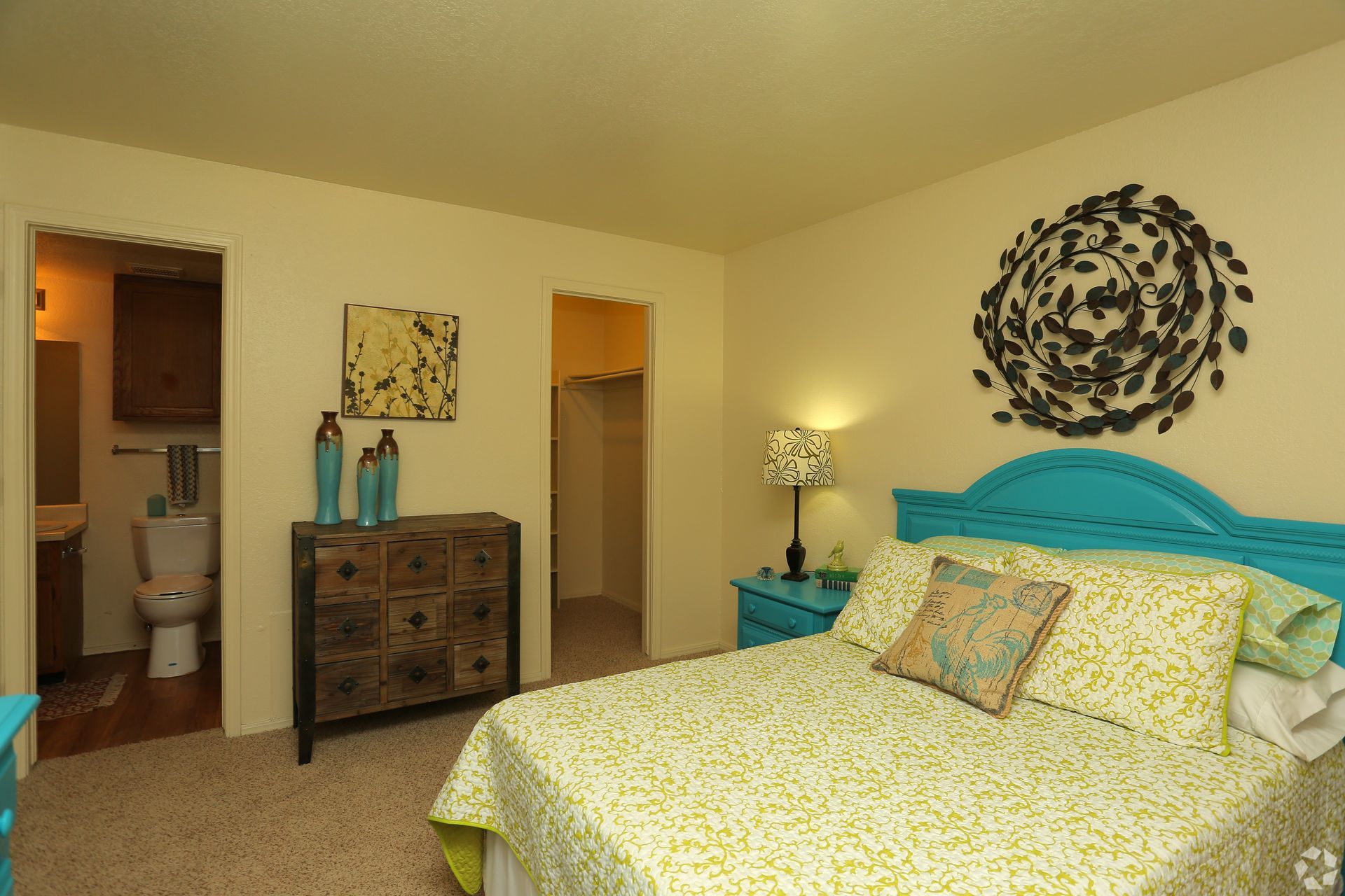 Bedroom with teal bed, patterned bedding, dresser, lamps, and decorative wall art.