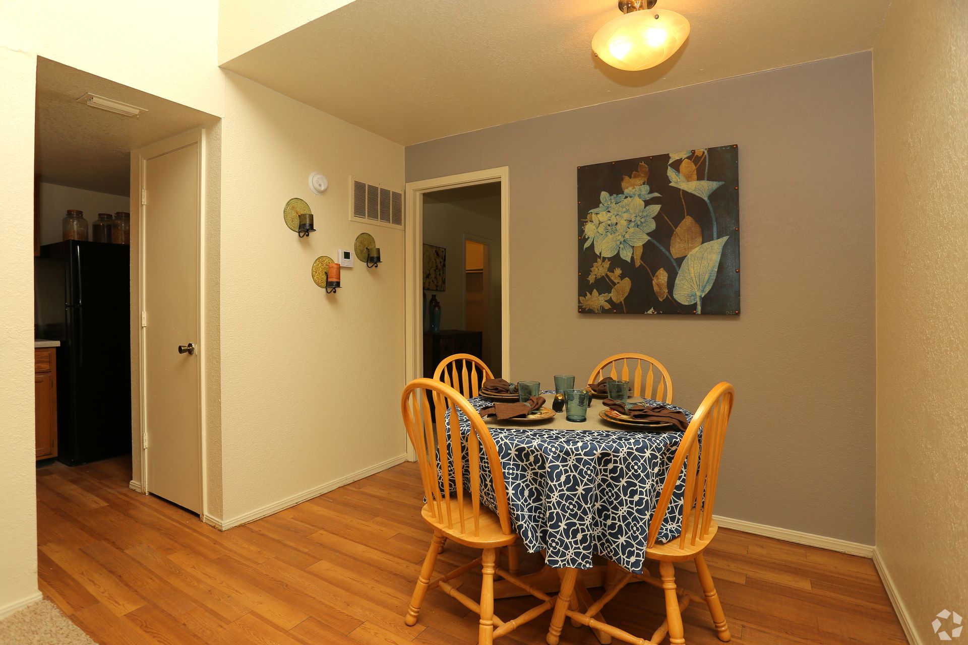 Dining room with a round table, four wooden chairs, blue patterned tablecloth, and wall art on gray walls.