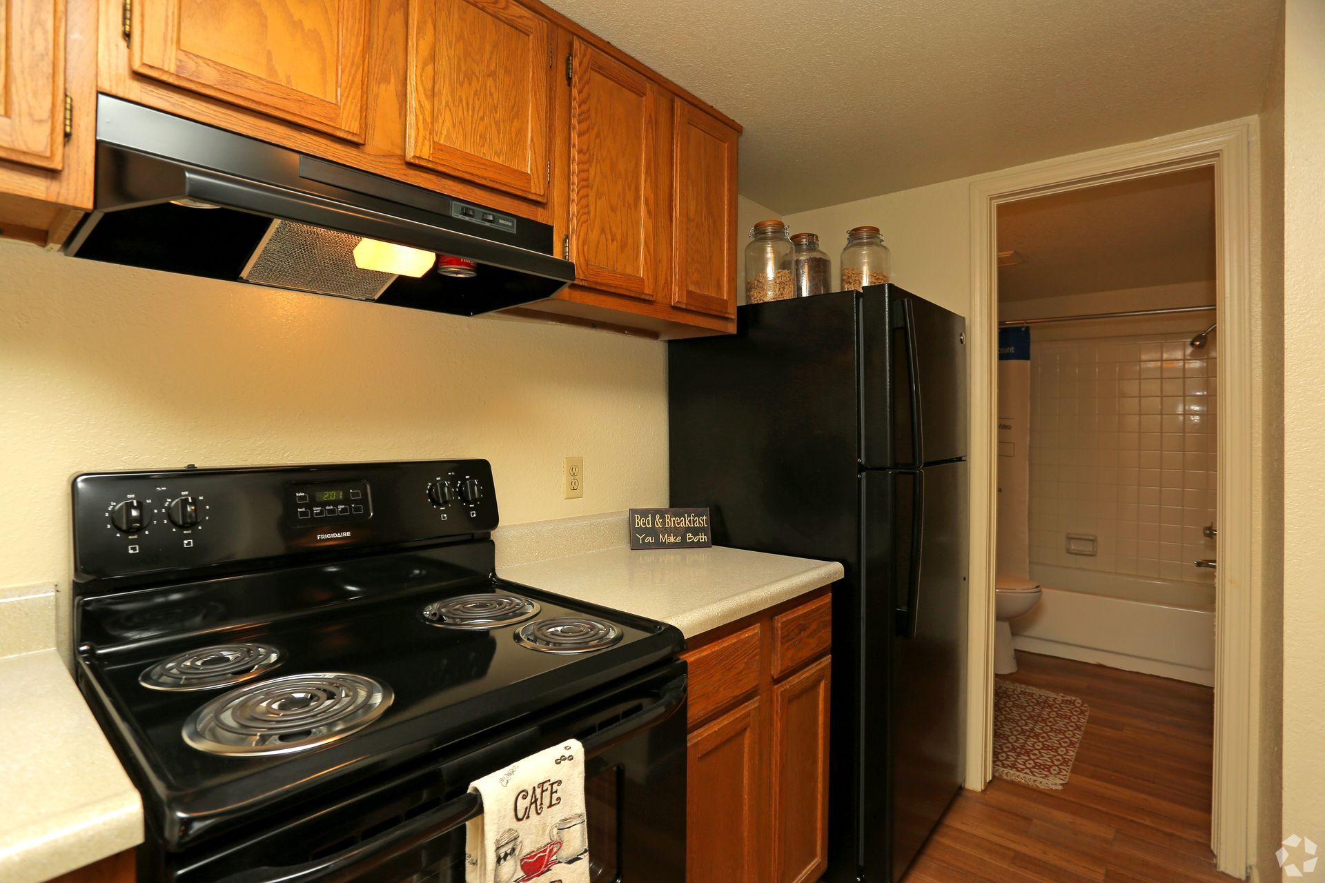 Small kitchen with black appliances, wooden cabinets, and a doorway to a tiled bathroom.
