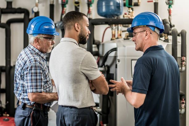 Three men wearing hard hats are standing in a room talking to each other.