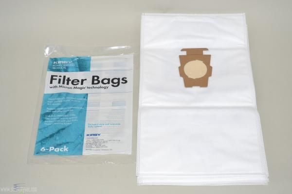 Pack of six vacuum cleaner filter bags, unopened, next to a stack of filter bags. White bags with brown filter cutout.