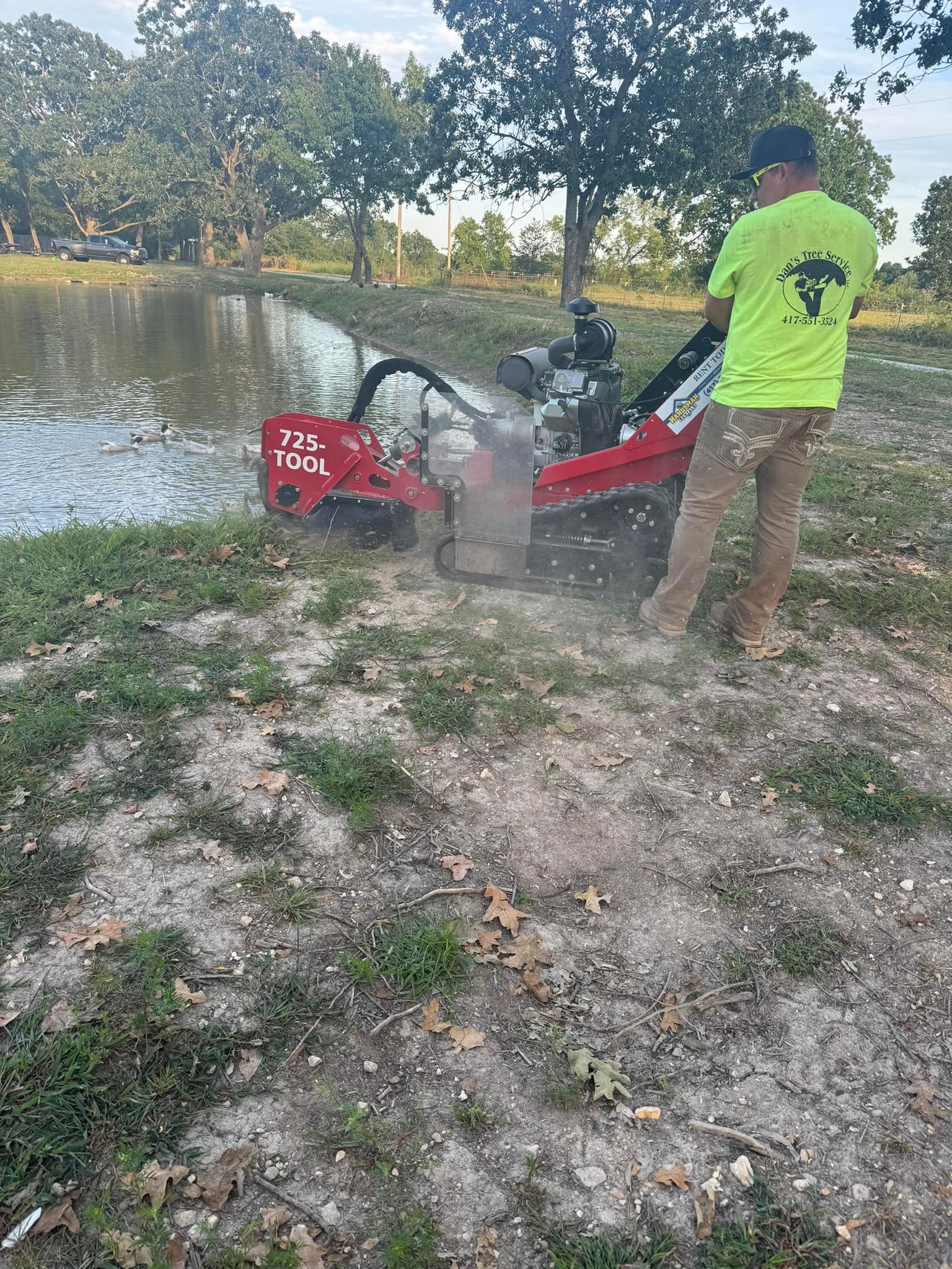 Man using a red machine to clear vegetation from a pond's edge. Green shirt, brown pants. Outdoors.