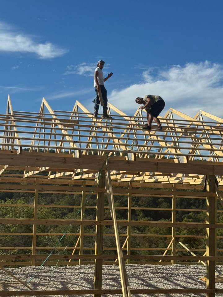Two people constructing a wooden building roof against a blue sky. One stands, the other works, both high up.