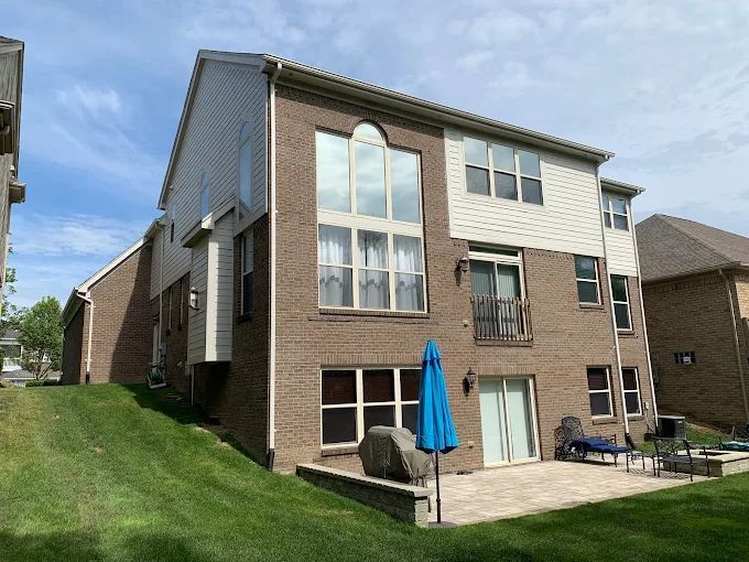 Back of a brick and white sided house on a sloped grassy yard with a patio and blue umbrella.