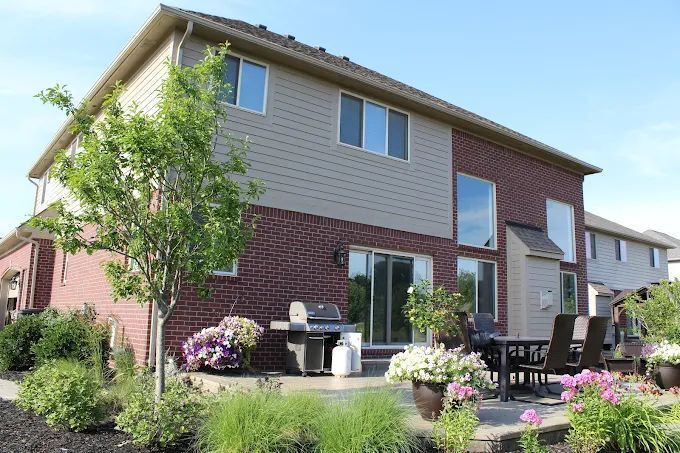 Back of a two-story house with brick and tan siding, patio, grill, and landscaped yard.