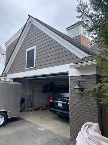 Gray-brown house with white trim, a partially open garage with a black SUV inside, and a trailer to the left.