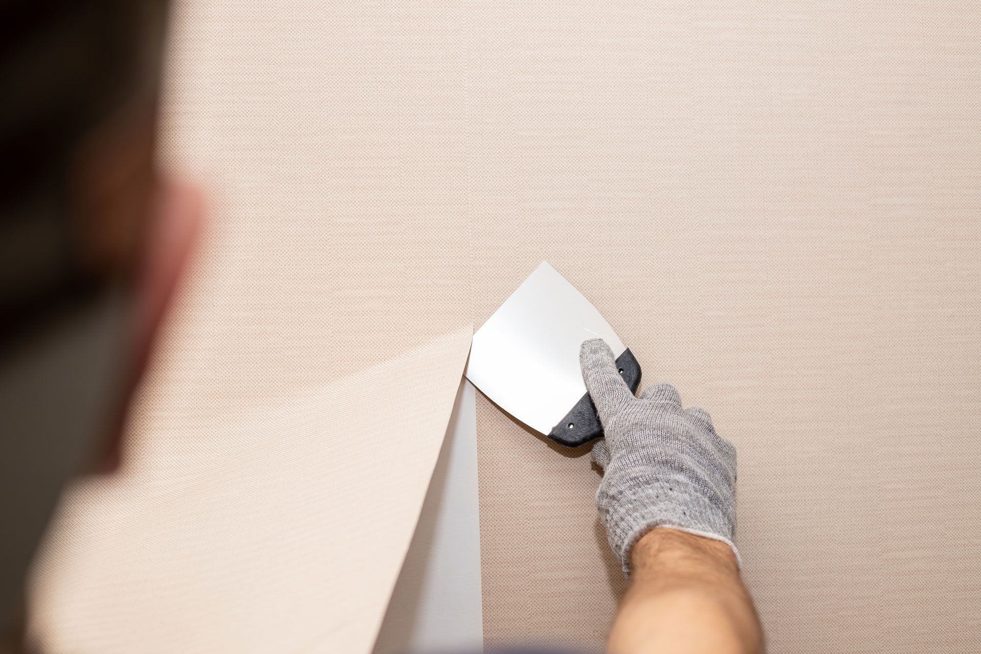 Person using a scraper to remove wallpaper from a wall, wearing a glove.