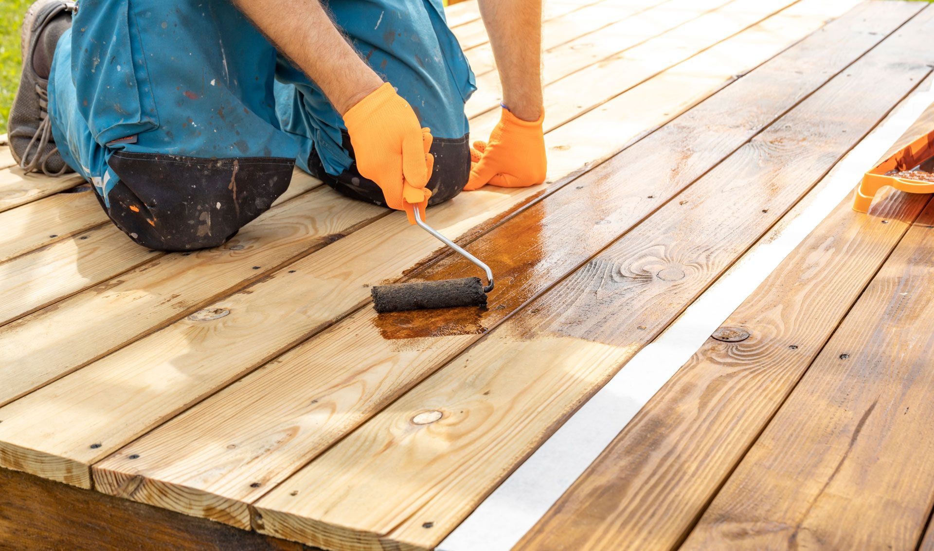 Person staining a wooden deck with a roller, wearing gloves and blue overalls.