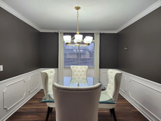 Dining room with white wainscoting, dark gray walls, glass table, and upholstered chairs. Golden chandelier hangs above.