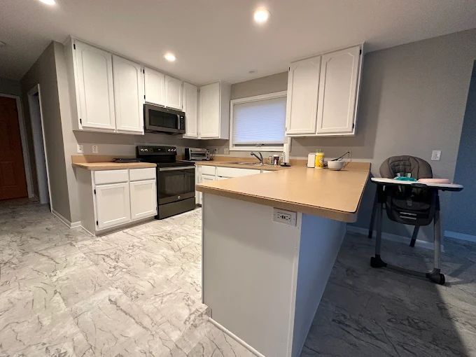 Kitchen with white cabinets, light countertops, black appliances, and grey flooring. A high chair sits to the side.