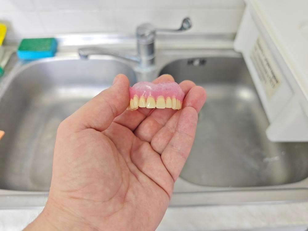 A Person Is Holding a Denture in Their Hand in Front of A Sink — Cosmetic Denture Services–Jacob Maxwell in Moranbah, QLD