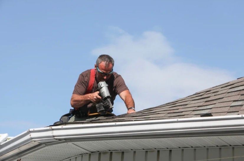 Worker using a power tool on a shingled roof under a blue sky