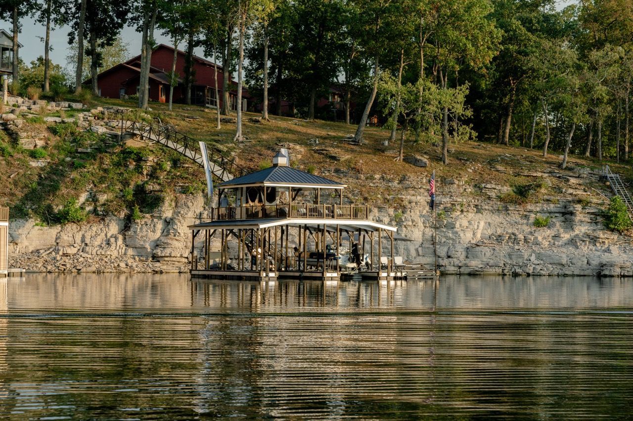 Wooden lakeside gazebo on a rocky shoreline beneath trees, reflected in calm water.