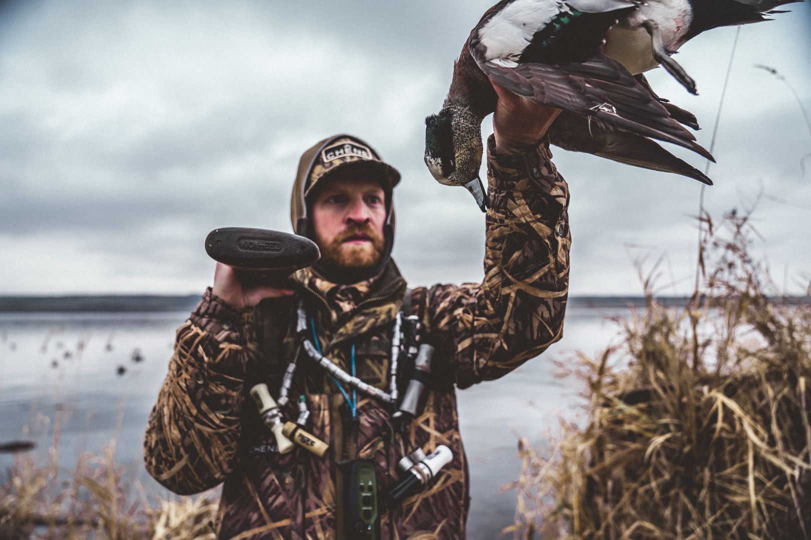 Hunter in camouflage holding a duck by a lakeshore under a cloudy sky