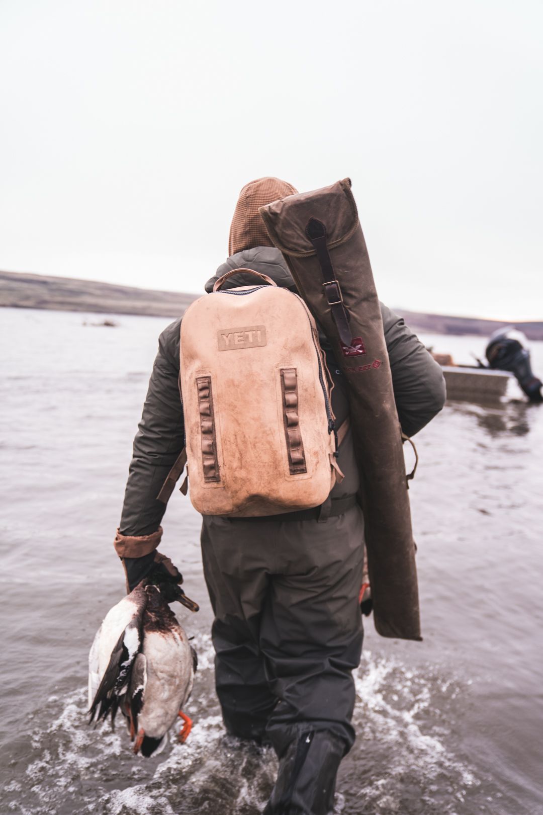 Person wades through shallow water carrying a backpack and a duck by the neck in a snowy shoreline setting