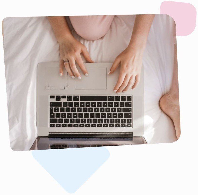 married woman dressed in pink sitting barefoot on her bed and using her laptop