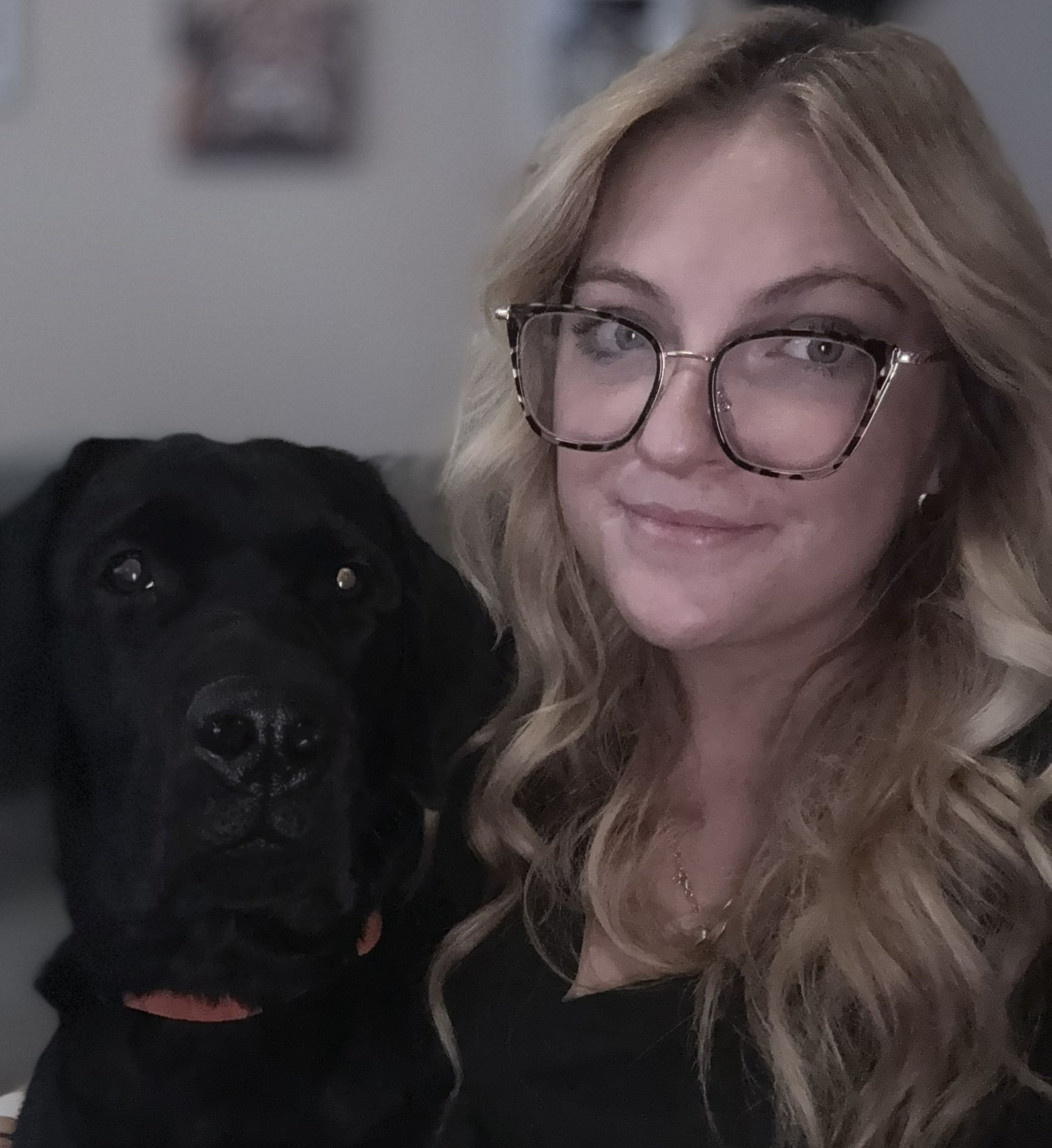 Woman with blonde hair and glasses sits next to a black Labrador.