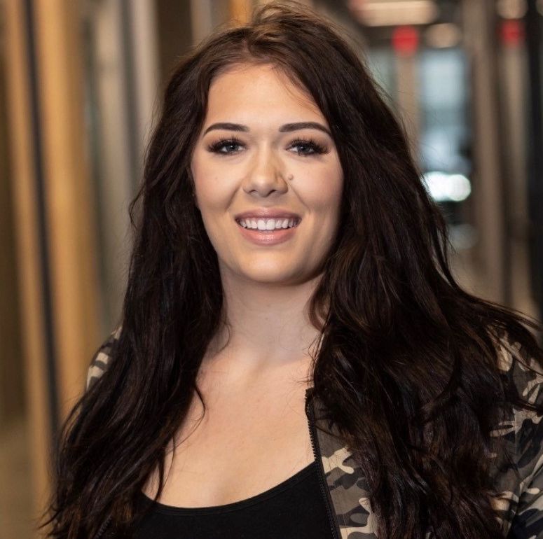 A woman with long dark hair is smiling and wearing a camouflage jacket.