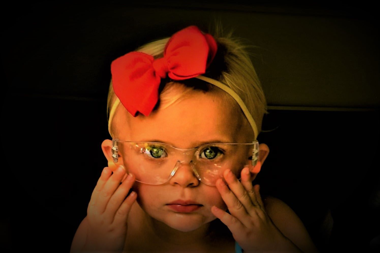Child with a red bow headband and glasses, hands on cheeks, looking intently.