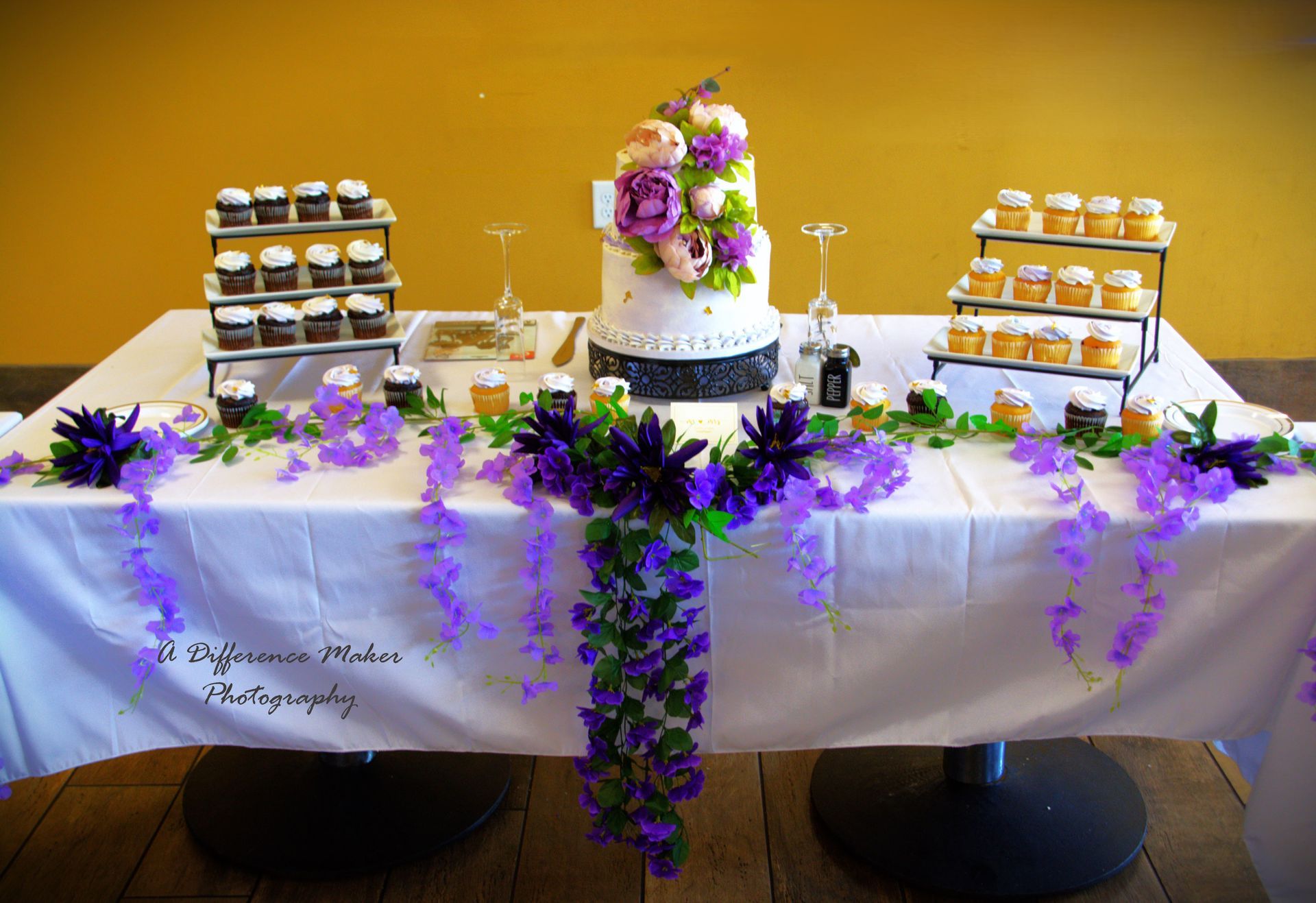 Wedding dessert table with cake, cupcakes, purple floral decorations on white tablecloth.