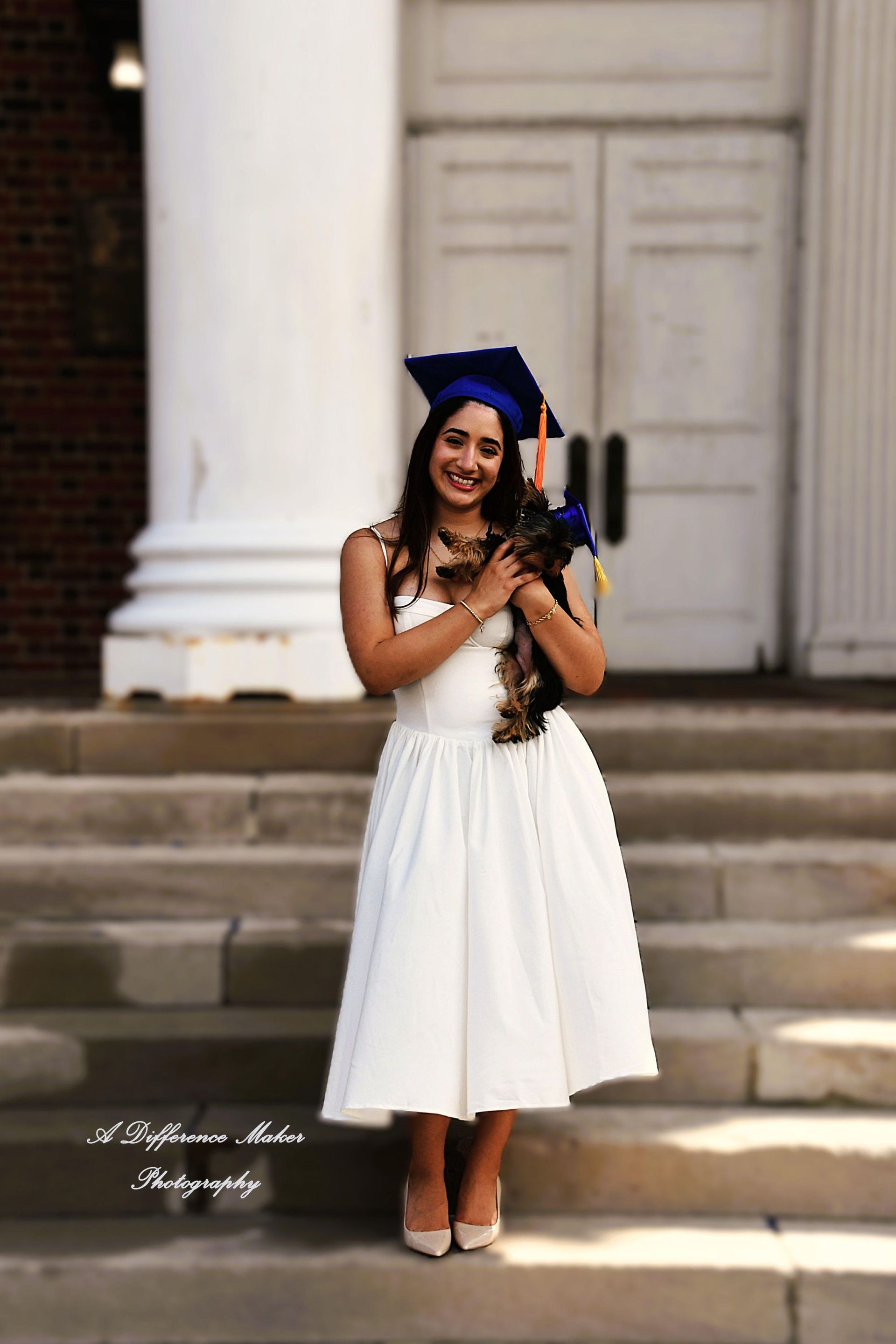 Woman in graduation attire holding a small dog on steps in front of a building.