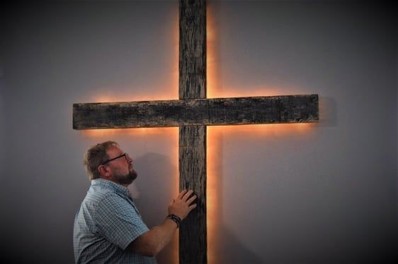Man touching large wooden cross on a wall with glowing light behind it.